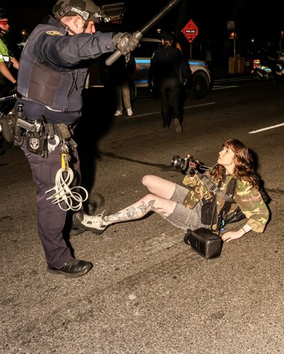 A police officer in riot gear swinging his baton over journalist Olga Fedorova after having pushed her to the ground, shortly before her arrest. Fedorova is staring defiantly back at the cop, still pointing her camera at him. Photo credit Alex Kent (alex-kent.com, via twitter @AlexKentTN)