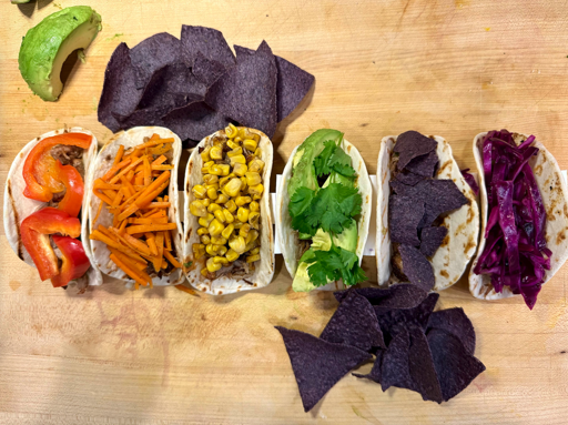 Six tacos arranged on a cutting board. Their topics include red bell pepper, orange carrot, yellow corn, green avocado, blue corn chips, and purple cabbage.