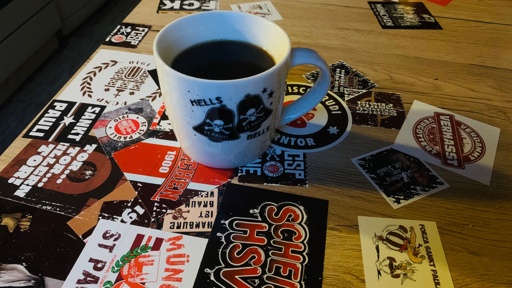 Auf einem Holztisch steht eine Kaffeetasse, die mit verschiedenen bunten Aufklebern, darunter Motive rund um St. Pauli und andere Fußballmotive, bedeckt ist. Die Tasse ist mit einem grafischen Design mit Totenköpfen und Glöckchen versehen.